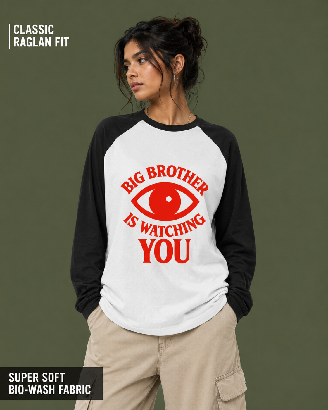 A woman wearing a raglan-style long-sleeve shirt with a white body and black sleeves, featuring a red graphic of an eye and the text 'BIG BROTHER IS WATCHING YOU' on the front. The background is a solid dark olive green.