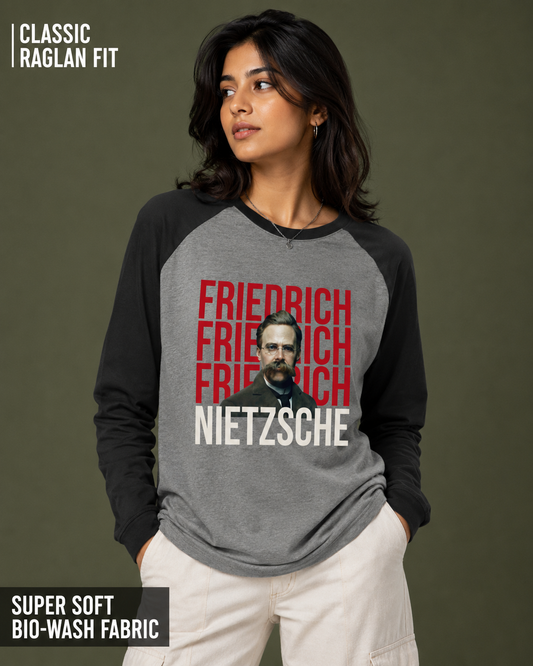 A woman modeling a raglan-style long-sleeve shirt with a heather gray body and black sleeves. The front features a graphic with repeated red 'FRIEDRICH' text, a black-and-white portrait, and white 'NIETZSCHE' text.